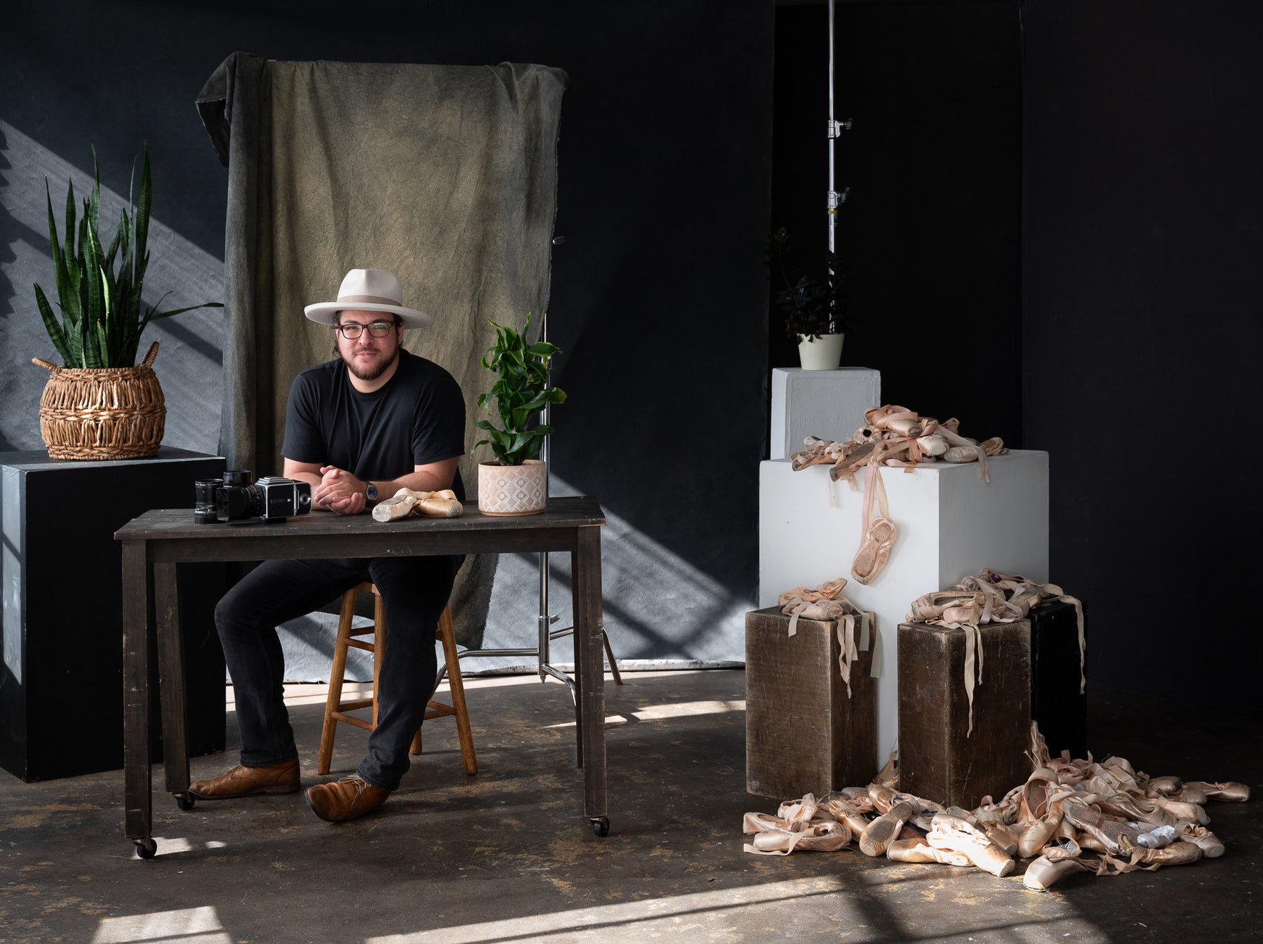 David Teran sits at a desk with pointe shoes, a hasselblad camera with a gold and black backdrop behind him