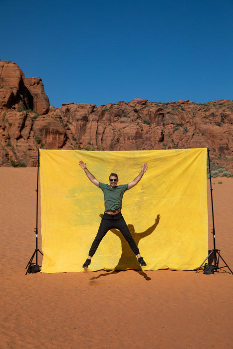 Matthew Peterson of Ultraviolet Backdrops walks in front of a giant, yellow, tie dye, pink, texturedmuslin backdrop while the sandstone cliffs of Snow Canyon loom in the background. Backdrop in a Bag are portable, lightweight solutions for on the go photographers. 