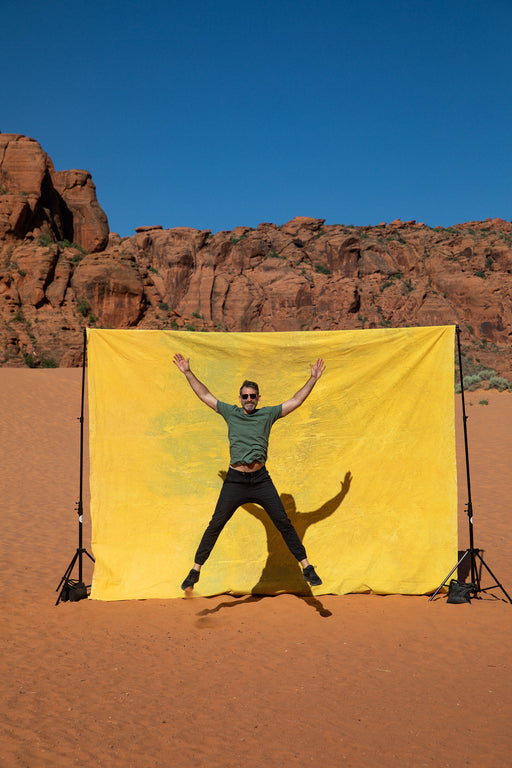 Matthew Peterson of Ultraviolet Backdrops walks in front of a giant, yellow, tie dye, pink, texturedmuslin backdrop while the sandstone cliffs of Snow Canyon loom in the background. Backdrop in a Bag are portable, lightweight solutions for on the go photographers.