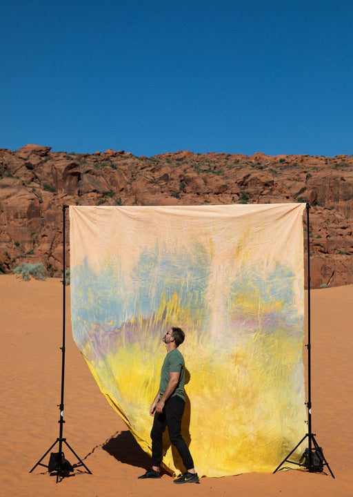 Matthew Peterson of Ultraviolet Backdrops walks in front of a giant, yellow, tie dye, pink, texturedmuslin backdrop while the sandstone cliffs of Snow Canyon loom in the background. Backdrop in a Bag are portable, lightweight solutions for on the go photographers. 
