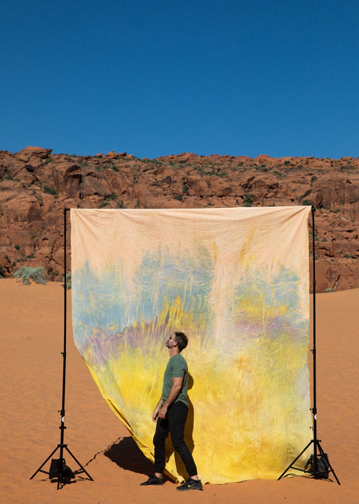 Matthew Peterson of Ultraviolet Backdrops walks in front of a giant, yellow, tie dye, pink, texturedmuslin backdrop while the sandstone cliffs of Snow Canyon loom in the background. Backdrop in a Bag are portable, lightweight solutions for on the go photographers.