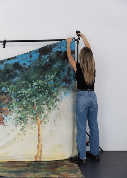 Person hanging a painted backdrop with a scenic design on a white wall.